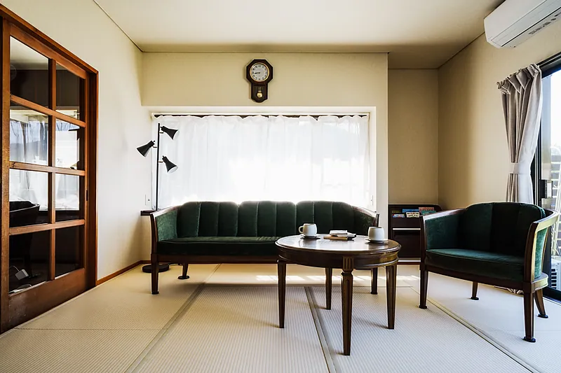 1階のリビング①の全景です。
当宿は全室「畳（日本伝統のフロア）」のお部屋ですが、床に座る文化のない海外のゲスト様にもゆっくりとくつろいでいただけるよう、このお部屋にはソファー（計4人掛け）をご用意いたしました。
ソファーはDREXEL HERITAGE、カフェ机はDomani（カリモク）、振り子時計はLANDEXで、いずれもアンティーク品です。
2つある間接照明を灯すと、これら家具と畳が調和し、より一層趣あるアンティークな和空間が演出されます。